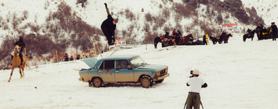 Person performing a snowboard trick over an old car in a snowy landscape with onlookers and photographers.