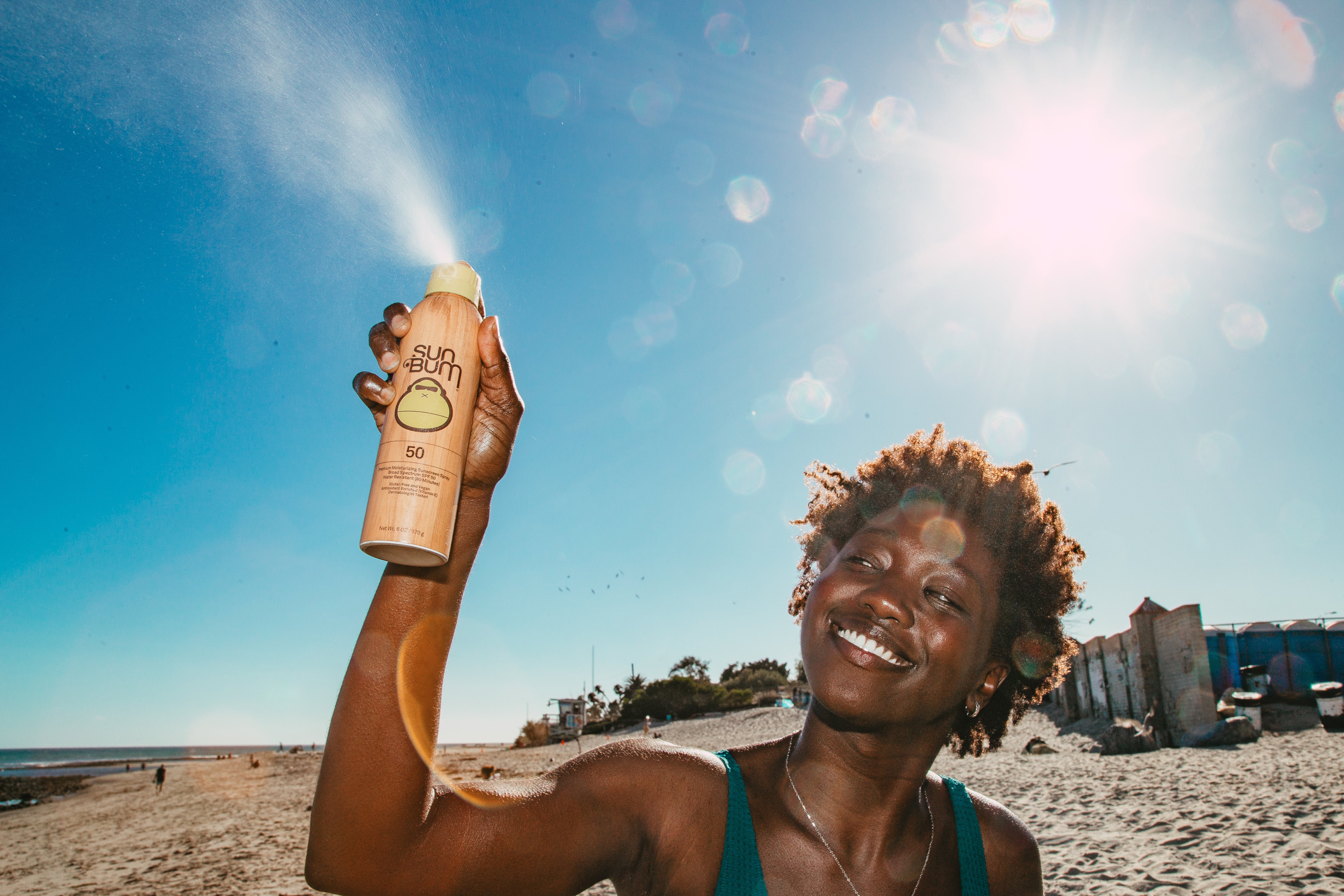 Woman holding a bottle of sunscreen on a beach with a clear blue sky.