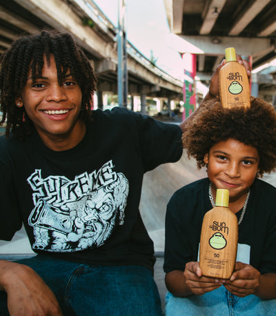 Two people holding Sun Bum sunscreen bottles under an overpass.